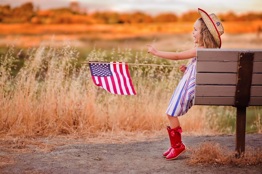 A young girl wearing red boots sitting on a bench holding an American Flag