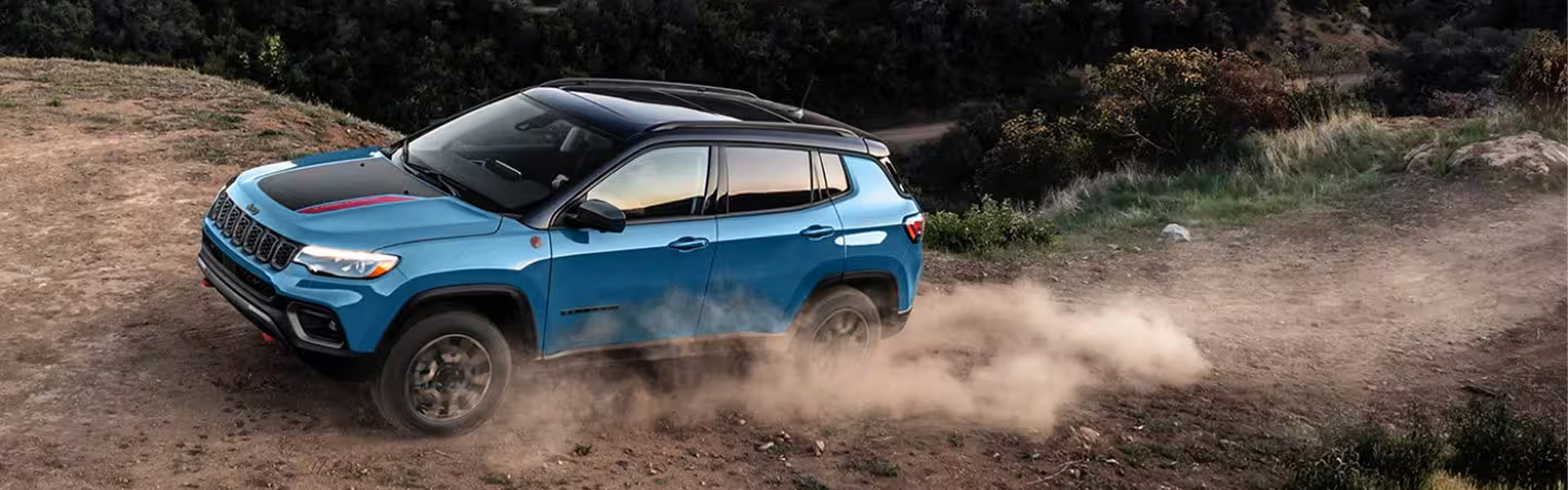 A teal blue 2025 Jeep Compass driving up a dirt hill with dust kicking out from behind the vehicle tires
